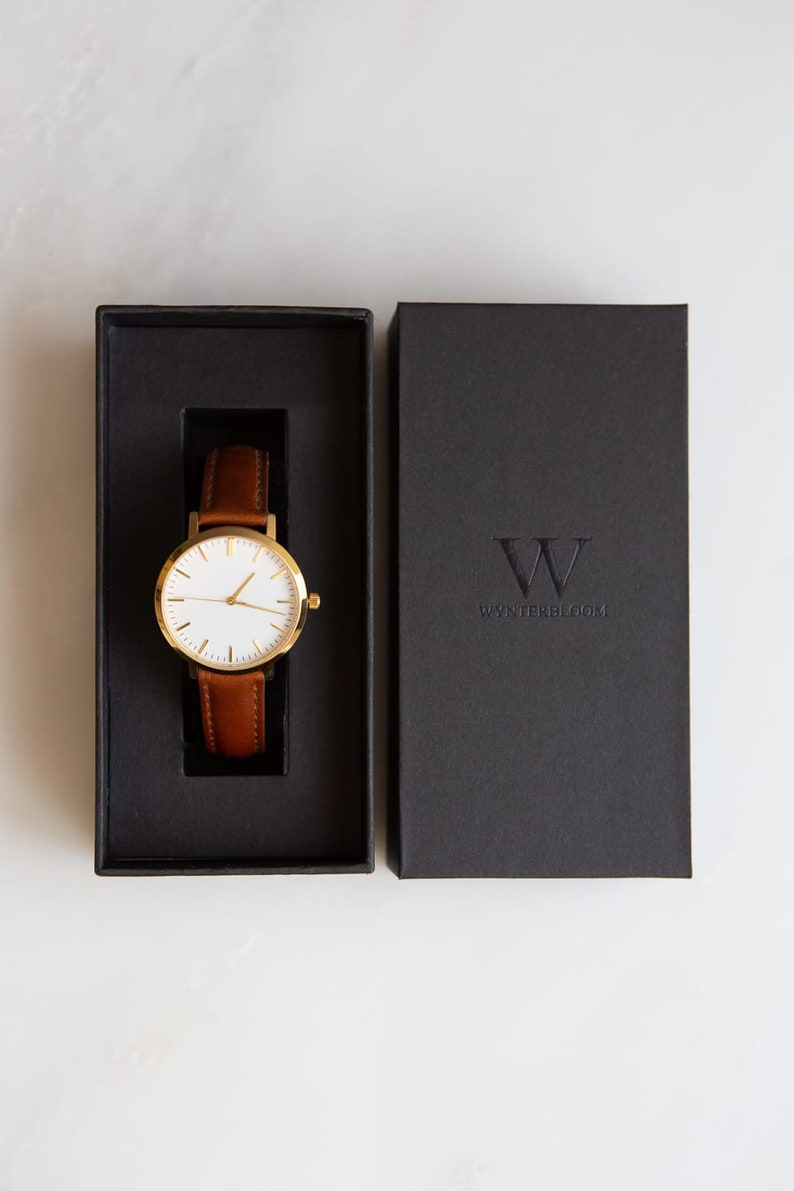 May include: A gold-tone wristwatch with a white face and brown leather band is displayed in an open black box. The watch is presented next to a closed black box with the embossed text "WYNTERBLOOM". The watch has a classic, minimalist design.