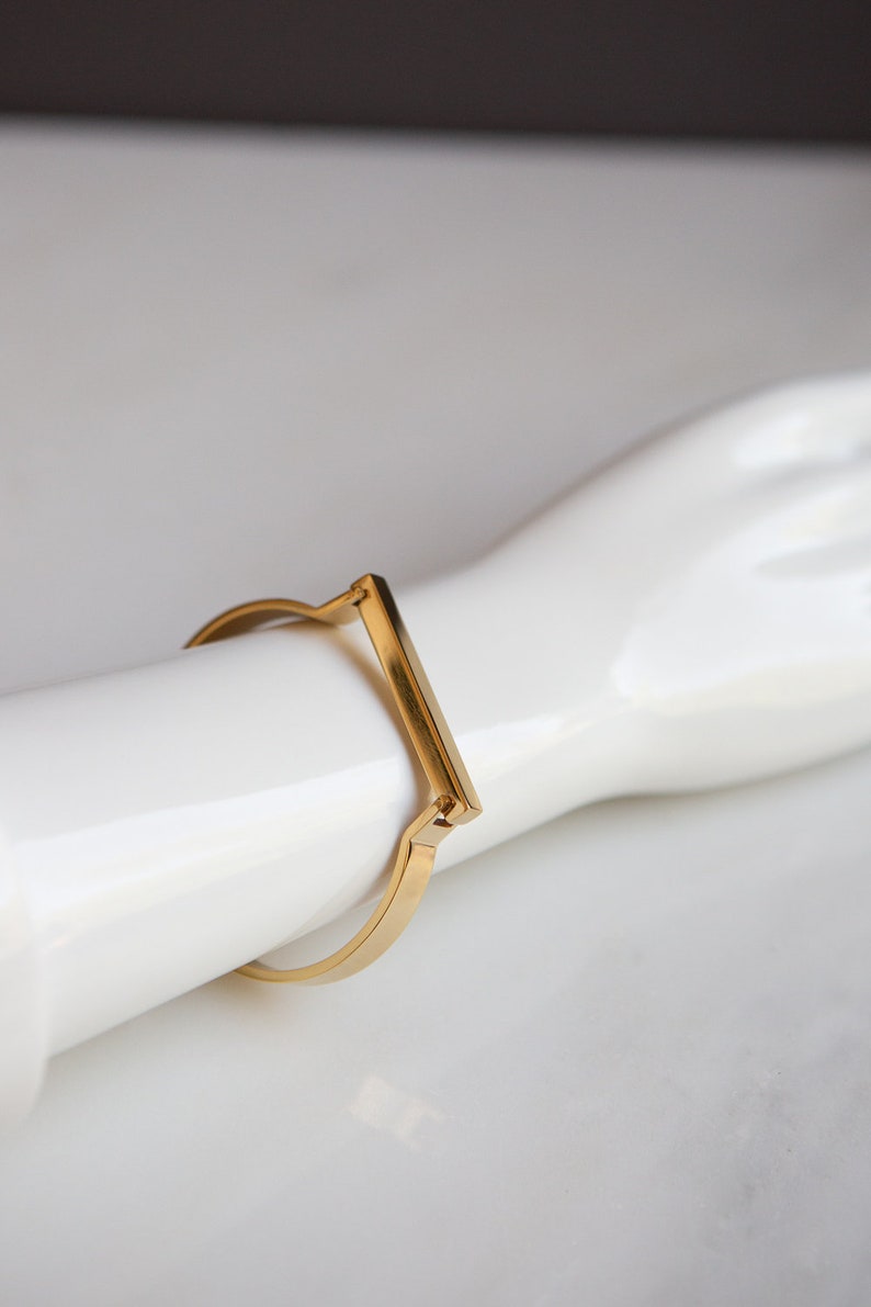 Op de afbeelding: Een gouden metalen armband met een rechthoekig balkontwerp.