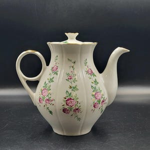 May include: A white porcelain teapot with a fluted, scalloped body and a matching lid. The teapot features a delicate floral pattern of pink roses and green leaves on each panel, accented with gold trim on the handle, spout, and lid rim.