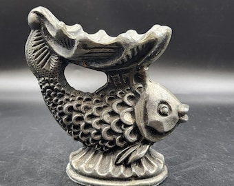 May include: A silver fish-shaped ashtray with a detailed design. The fish has scales and a fin on its back. The ashtray is a decorative piece for a home or office.