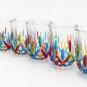 Peut inclure: Un ensemble de six verres transparents disposés en rangée. Chaque verre présente un bord évasé et une base ornée d'un motif multicolore vibrant composé de formes peintes en rouge, bleu, jaune et vert rappelant des pétales de fleurs.