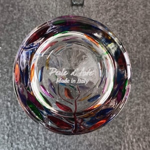 May include: The bottom of a clear glass tumbler with a colorful design of leaves and dots. The glass is marked with the text "Pelle d'Arte Made in Italy".