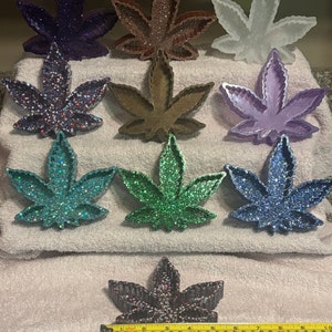 May include: Nine different colored resin leaf-shaped trinket dishes. The dishes are decorated with glitter and are arranged in three rows on a white towel. A measuring tape is visible at the bottom of the image.