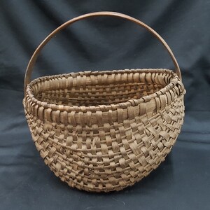 May include: A large, brown, woven basket with a wooden handle. The basket is made of natural materials and has a rustic, vintage look.