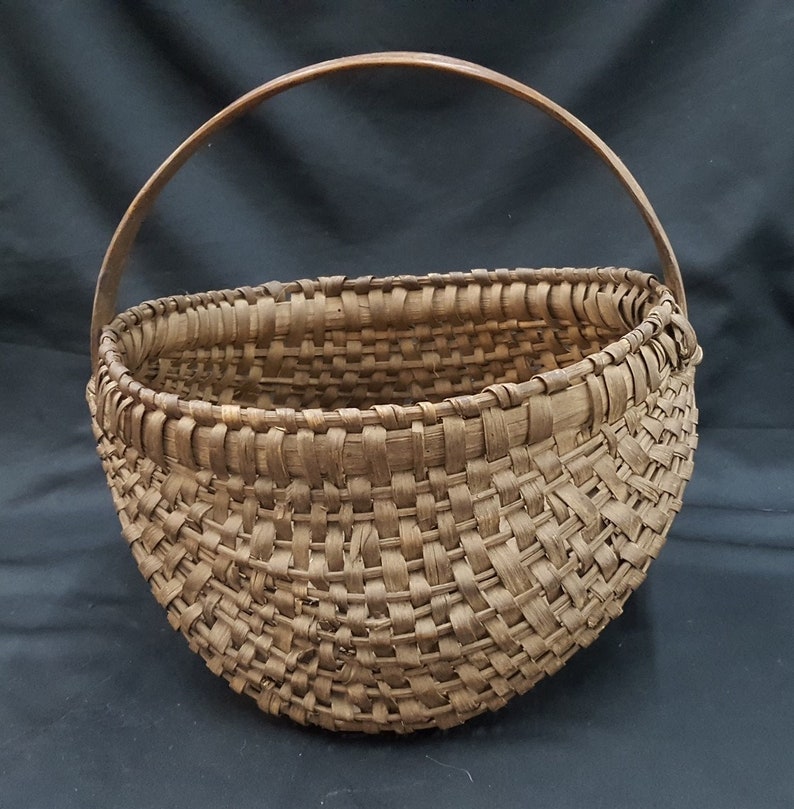 May include: A large, brown, woven basket with a wooden handle. The basket is made of natural materials and has a rustic, vintage look.