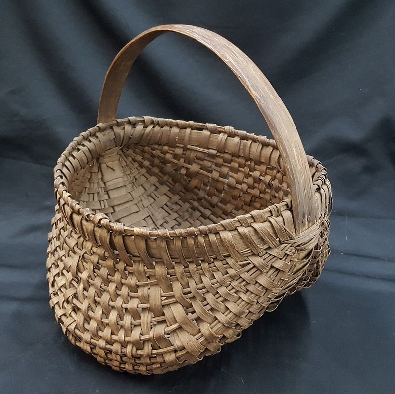 May include: A woven brown basket with a wooden handle. The basket is oval shaped and has a wide opening.