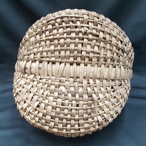 May include: A woven basket made of natural materials, with a rounded shape and a tightly woven pattern. The basket is light brown in color and has a smooth, even texture.