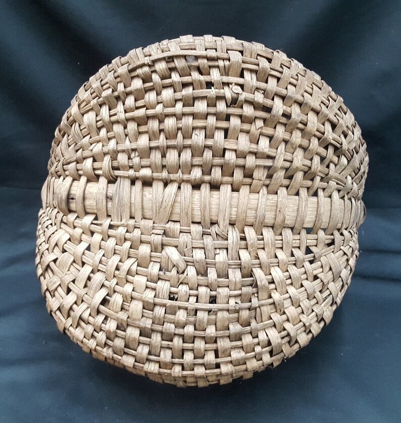 May include: A woven basket made of natural materials, with a rounded shape and a tightly woven pattern. The basket is light brown in color and has a smooth, even texture.