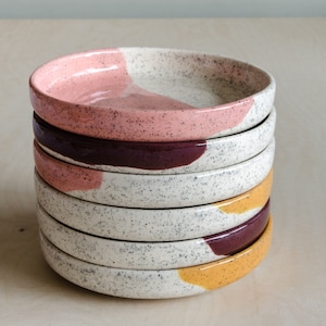 May include: A stack of six ceramic plates with speckled white glaze and colourful abstract designs in pink, burgundy, and yellow.