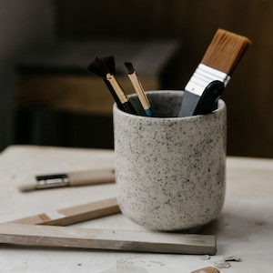 Op de afbeelding: Een gespikkelde keramische beker gevuld met kwasten, waaronder een waaierkwast, een ronde kwast en een platte kwast. De beker staat op een wit oppervlak met een houten liniaal en een kleine houten stempel.