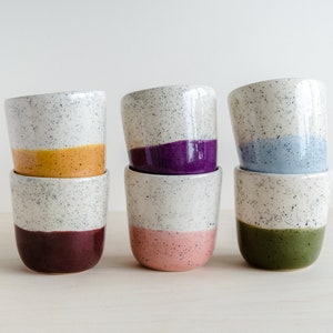 May include: Set of six speckled ceramic mugs with different colored bands. The mugs are stacked in pairs, with the top mug of each pair showing a different color band: yellow, purple, blue, and green.
