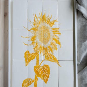 May include: A white rectangular frame containing twelve square tiles arranged in a grid. The tiles feature a yellow sunflower illustration with detailed petals and leaves, spanning across the individual tiles to form a single cohesive image.