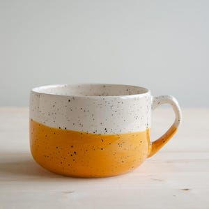 Könnte beinhalten: Ein breiter Keramikbecher mit zweifarbigem Design, bestehend aus einem gesprenkelten weißen oberen Teil und einem einfarbigen senfgelben unteren Teil. Der Becher hat einen einzelnen geschwungenen Henkel an der rechten Seite und eine glatte Oberfläche.