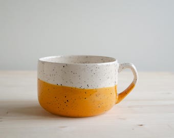 May include: A wide ceramic mug featuring a two-tone design with a speckled white upper section and a solid mustard yellow lower section. The mug has a single curved handle on the right side and a smooth, glossy finish throughout the exterior surface.