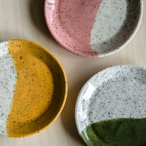 Könnte beinhalten: Drei Keramikteller mit gesprenkeltem weißen Überzug und einem kontrastierenden, halbkreisförmigen Design. Die Teller sind rosa und weiß, gelb und weiß sowie grün und weiß.