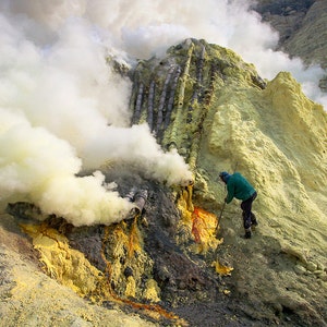 Puede incluir: Una persona con una chaqueta verde recoge azufre de una ventilación volcánica. La ventilación emite humo blanco y el área circundante está cubierta de depósitos de azufre amarillo.