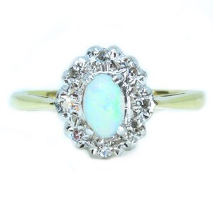 May include: An oval opal gemstone ring with a halo of small diamonds. The ring features a gold band and a cluster of diamonds surrounding the opal. The opal has a pale blue-green color.