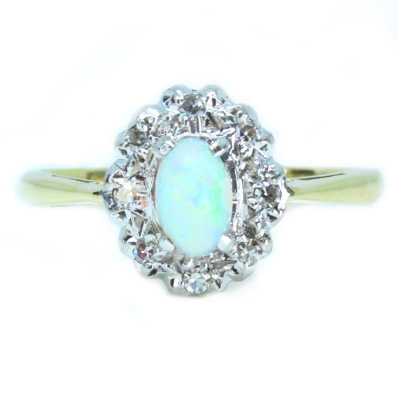 May include: An oval opal gemstone ring with a halo of small diamonds. The ring features a gold band and a cluster of diamonds surrounding the opal. The opal has a pale blue-green color.