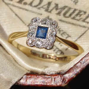 May include: A gold ring featuring a central square-cut blue sapphire surrounded by a scalloped halo of small round diamonds. The setting is crafted in a bright metal, contrasting with the polished gold band that tapers toward the center stone.