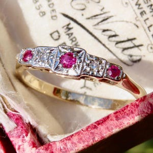 Peut inclure: Une bague en or avec une série de sertissages carrés et rectangulaires. Chaque sertissage contient une petite pierre précieuse rose vif et ronde. La bague est posée sur un fond rose tendre.