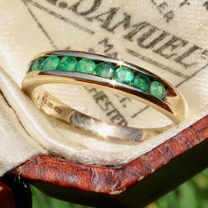 May include: A gold band ring featuring a channel setting with a row of seven round-cut green emerald gemstones. The interior of the band displays visible hallmarks and engravings, showcasing a polished metallic finish and a classic circular design.