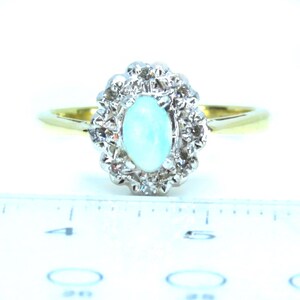 May include: A gold band ring with a central oval, light blue opal stone, surrounded by a halo of small, clear gemstones. The ring is set against a white background, showcasing its elegant design.
