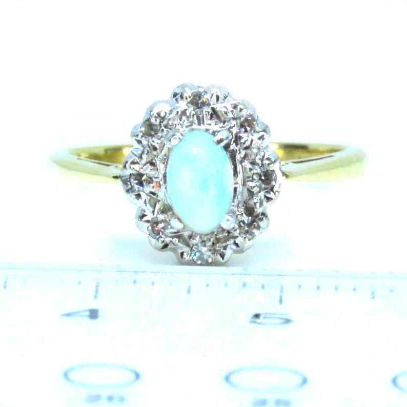 May include: A gold band ring with a central oval, light blue opal stone, surrounded by a halo of small, clear gemstones. The ring is set against a white background, showcasing its elegant design.