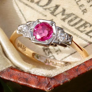 May include: A gold ring featuring a central round pink gemstone, with smaller clear stones on either side. The ring is set in a detailed, vintage style and is presented in a jewellery box. The ring band has the number "19335" engraved.