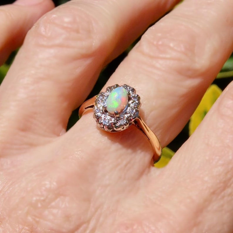 May include: A gold ring featuring an oval opal gemstone surrounded by a halo of small, clear gemstones. The opal displays a play of color with hints of green, pink, and blue. The ring is set on a slender gold band.