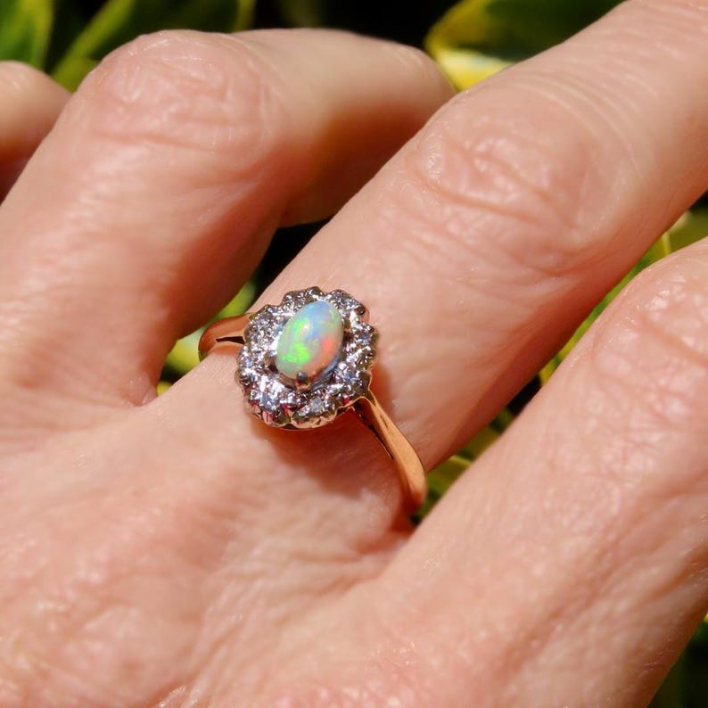 May include: A gold ring featuring an oval opal gemstone surrounded by a halo of small, clear gemstones. The opal displays a play of color with hints of green, blue, and yellow. The ring is set on a delicate gold band.