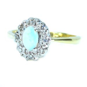 May include: An oval opal gemstone ring with a halo of small diamonds set in a silver-colored metal. The ring's band is gold-colored. The opal has a pale blue-green hue.