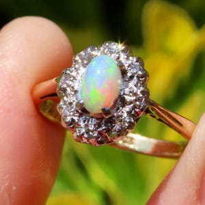 May include: A gold ring featuring an oval opal gemstone with a halo of small, sparkling stones. The opal displays a play of color with hints of green, pink, and blue. The ring is held between fingers against a blurred background.