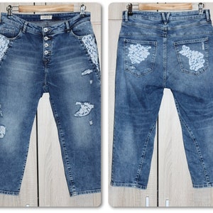 Pode incluir: Um par de calças de ganga azuis com detalhes de renda branca nos bolsos da frente e de trás. As calças têm um aspeto desgastado com rasgos e rasgões por todo o tecido.