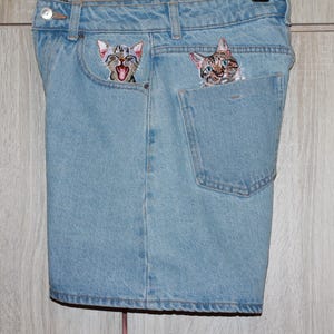 Pode incluir: Um par de calções de ganga azul claro com dois rostos de gato bordados a espreitar das alças. O gato à esquerda está a olhar para a direita e o gato à direita está a olhar para a esquerda.