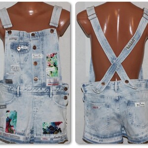 Könnte beinhalten: Hellblaue Jeans-Latzhosen mit Used-Look und verschiedenen Patches und Texten. Die Latzhosen haben verstellbare Träger und einen Knopfverschluss.
