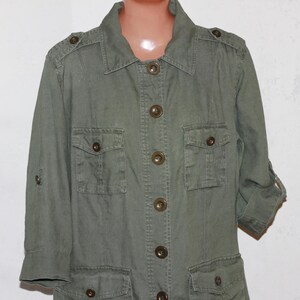 Op de afbeelding: Olijfgroene utility jas met vier voorzakken, knoopsluitingen en oprolbare mouwen. De jas heeft een kraag en epauletten op de schouders. De knopen zijn donkerbruin.