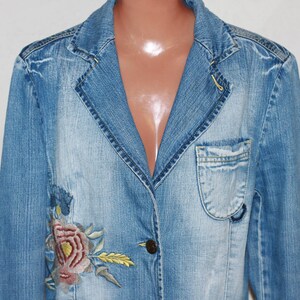 Peut inclure: Une veste en jean bleu clair avec un motif de broderie florale sur le côté gauche. La veste a un col classique, une fermeture à boutons et deux poches sur le devant.