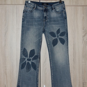 May include: Light blue denim jeans with a floral design. The jeans have a classic five-pocket design and a straight leg. The floral design is a darker shade of blue and is placed on the front of the jeans.