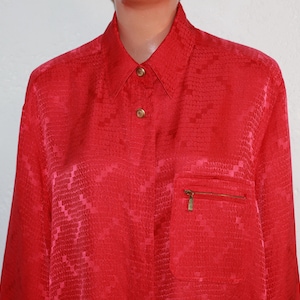 May include: A vibrant red button-down shirt with a textured pattern. The shirt features a collared neckline, gold-colored buttons, and a pocket with a gold zipper. The fabric has a subtle sheen.