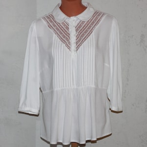 May include: White blouse with a Peter Pan collar and three-quarter sleeves. The bodice features vertical pleats and a decorative lace panel in a muted pink hue. The blouse has a gathered waist and a relaxed fit.