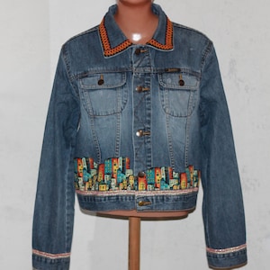 May include: A denim jacket with a cityscape design. The jacket has a light blue wash, an orange decorative collar, and a colourful cityscape design along the bottom. The cuffs and bottom hem have a decorative trim.