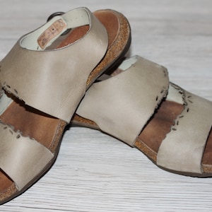 May include: A pair of beige leather sandals with cork soles and decorative stitching. The sandals have two straps across the foot and an ankle strap.