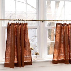 May include: Two brown linen cafe curtains hang from a gold curtain rod in front of a window. The curtains are gathered at the top and have a simple, rustic look.