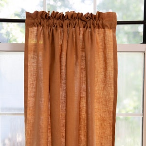 May include: A pair of rust-colored linen kitchen valances with a gathered top and a simple hem. The valances are hanging in a window.