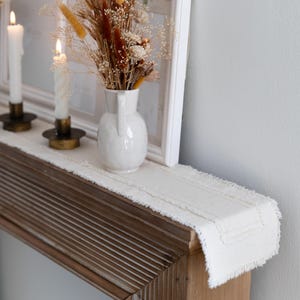 May include: A decorative display featuring a white vase with dried flowers, lit candles in brass holders, and a white framed picture. A cream-colored fringed table runner sits on a wooden surface.