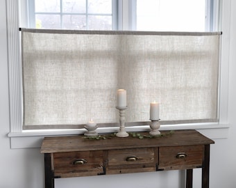 May include: A neutral-toned linen window treatment hangs above a rustic wooden console table. The table features three drawers with brass handles. Lit candles in decorative holders and greenery add a warm ambiance to the scene.