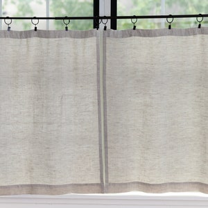 May include: A pair of off-white linen curtains with a subtle texture hang from a black curtain rod. The curtains have a thin, darker gray trim at the bottom.