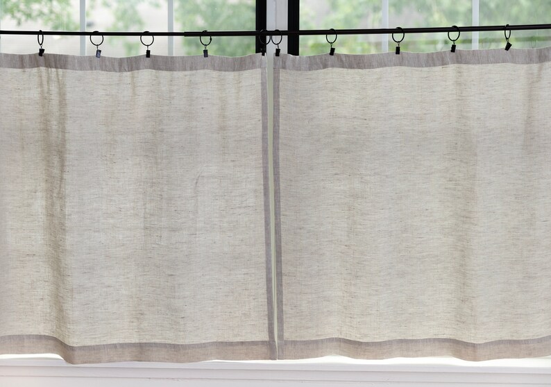 May include: A pair of off-white linen curtains with a subtle texture hang from a black curtain rod. The curtains have a thin, darker gray trim at the bottom.