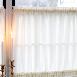 May include: Two white sheer cafe curtains with a ruffled edge hang in a window. Two lit candles are in gold candle holders in front of the curtains.