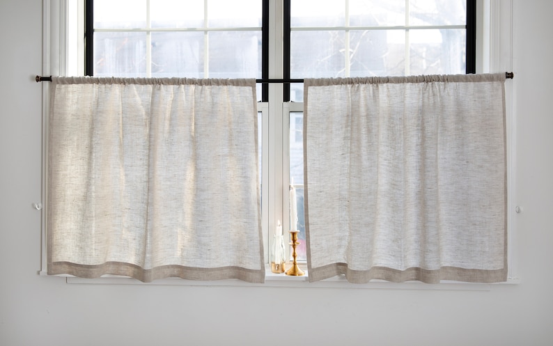 May include: A pair of white linen cafe curtains with a brown trim hang on a window with a black rod. Two gold candle holders with white candles are on the window sill.